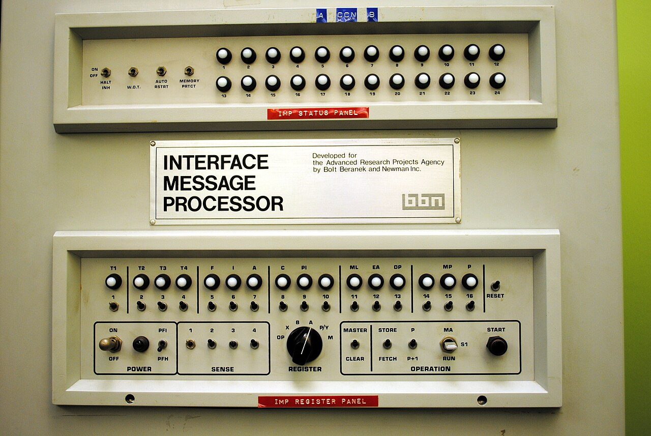 Painel frontal bege de uma máquina antiga, com fileiras de botões redondos e chaves, e uma placa no meio com o nome Interface Message Processor.