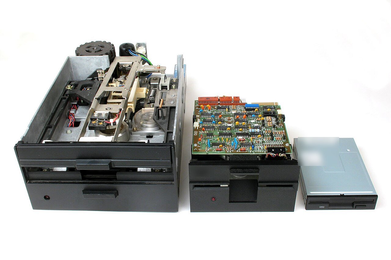Três leitores de disquete lado a lado sobre fundo branco, do maior para o menor: um leitor grande e aberto, mostrando a mecânica, um médio com uma placa de circuito em cima e um pequeno, fino e cinza.